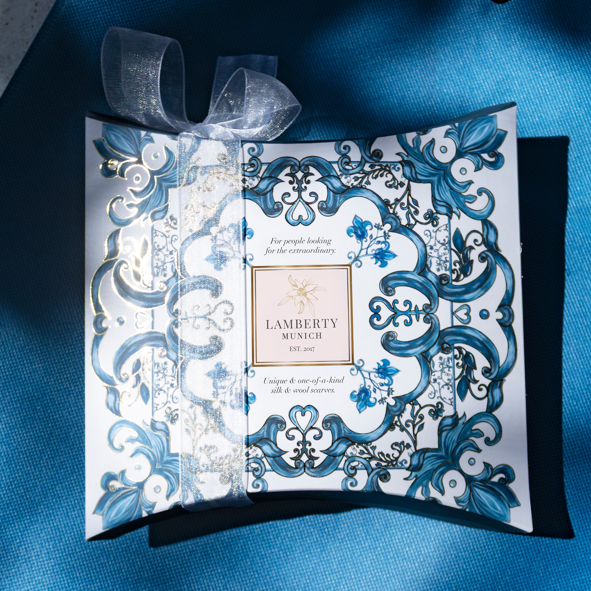 Lamberty Munich gift box with a blue and white Majolica pattern, a gold-accented brand label and a sheer organza ribbon, resting on blue fabric